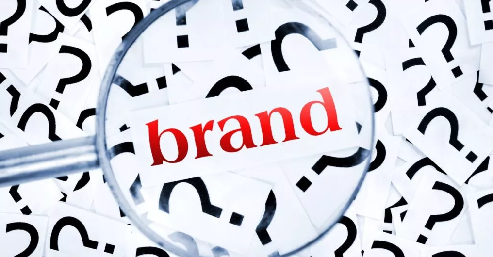 This is a stock image of the word "brand", shown through a magnifying glass, found in a pile of question marks.